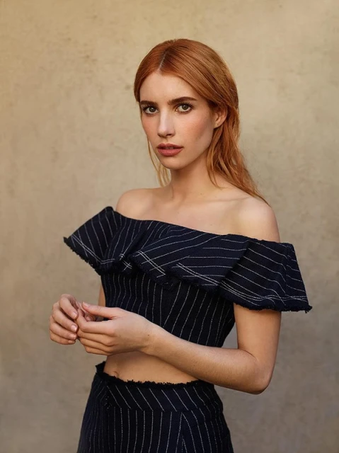 Emma Roberts portrait: A fair-skinned woman with auburn hair poses in a striped, off-the-shoulder top against a neutral backdrop.