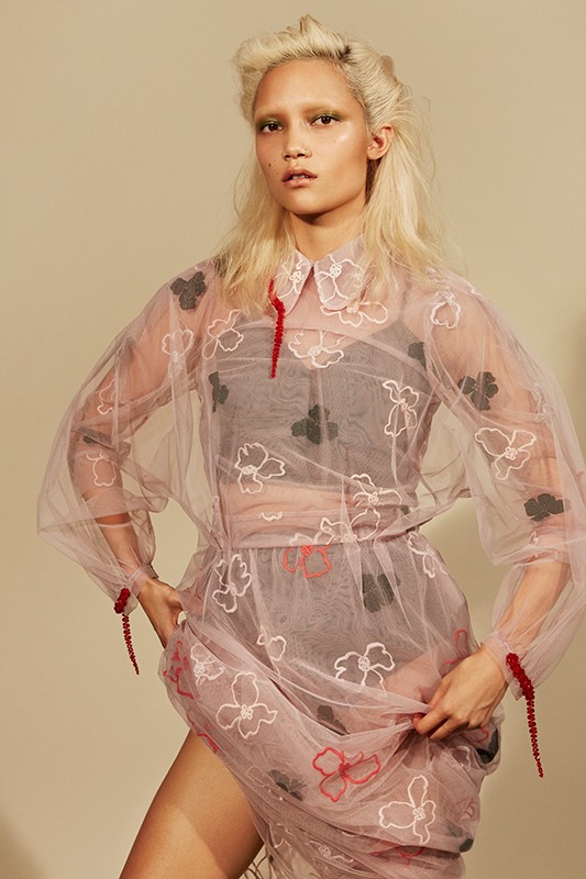 A stylish woman poses in a sheer floral dress with matching makeup and platinum hair against a neutral background, looking at the camera.