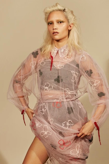 A stylish woman poses in a sheer floral dress with matching makeup and platinum hair against a neutral background, looking at the camera.
