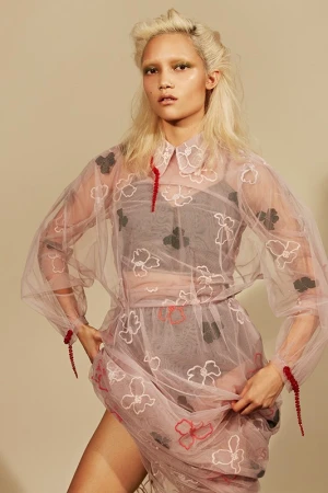 A stylish woman poses in a sheer floral dress with matching makeup and platinum hair against a neutral background, looking at the camera.