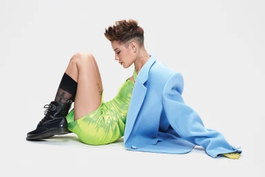 Woman with short textured hair sitting on a white seamless background wearing a lime green dress and a light blue blazer draped over her shoulder