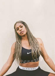 Black woman with long blonde-highlighted box braids wearing a black sports bra and shorts against a textured textured wall.