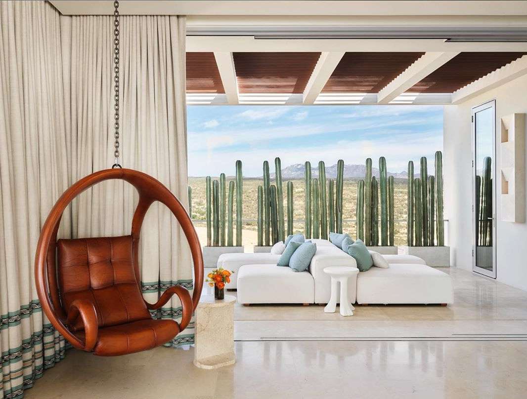 Modern interior with a hanging chair, modular sofa, and view of desert landscape with cacti, showcasing contemporary design and relaxation.