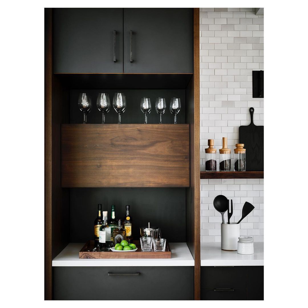Interior view of a modern kitchen bar area with wine glasses, liquor bottles, limes, and bar tools on white countertops.