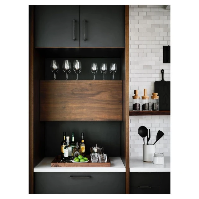 Interior view of a modern kitchen bar area with wine glasses, liquor bottles, limes, and bar tools on white countertops.