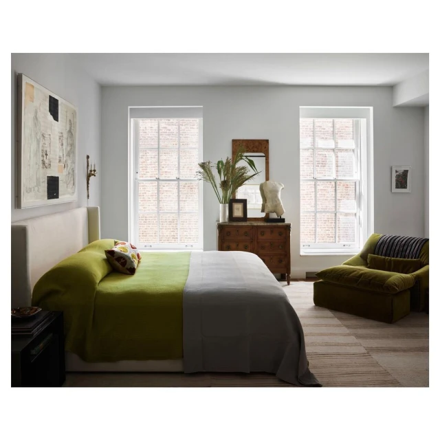 Bedroom interior with a green and grey bed, wooden dresser, windows, art, olive-green chair, and neutral tones in a naturally lit setting.