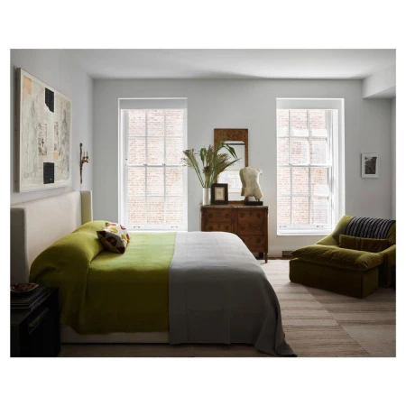 Bedroom interior with a green and grey bed, wooden dresser, windows, art, olive-green chair, and neutral tones in a naturally lit setting.