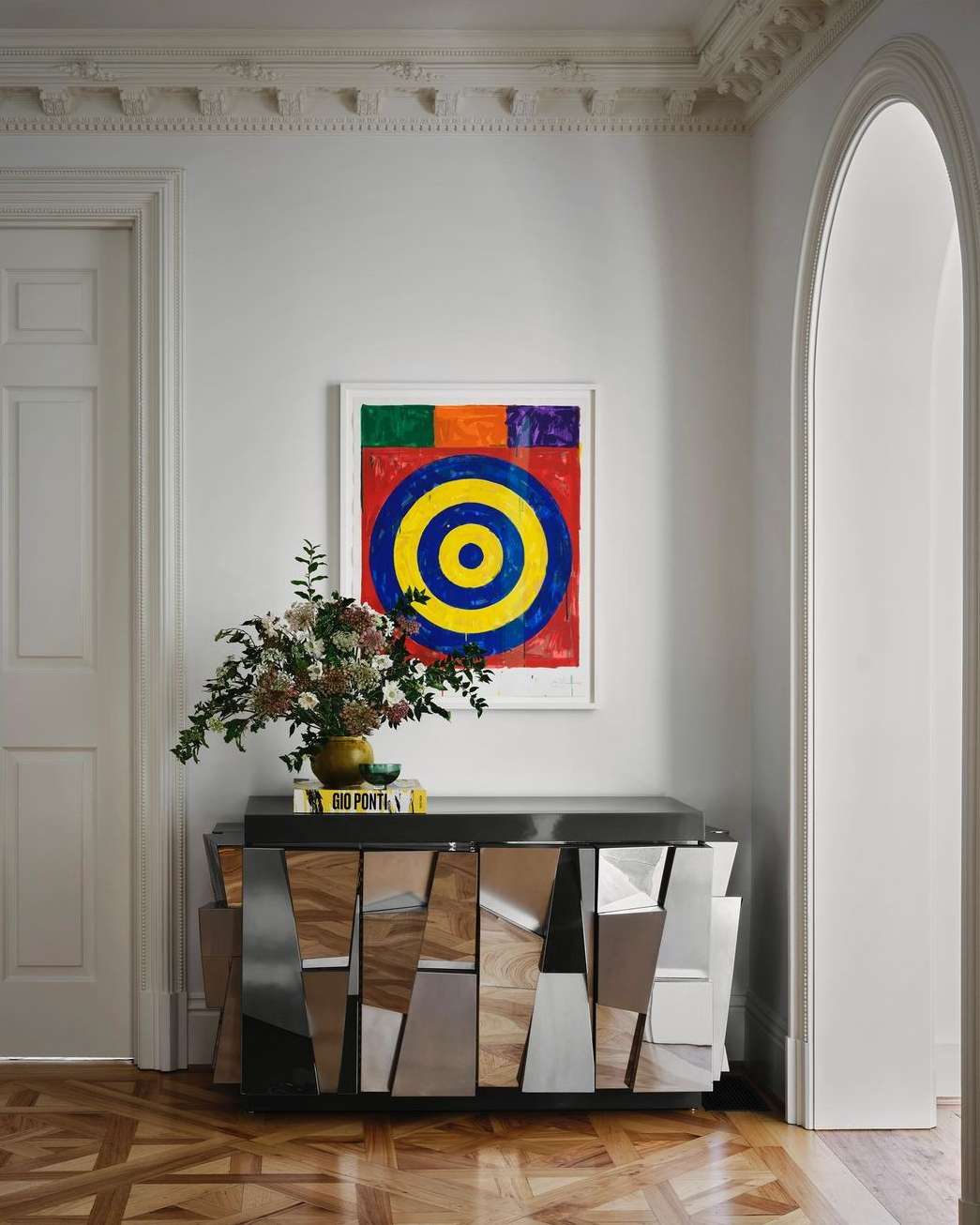 Interior design shot featuring a wooden cabinet with mirrored details, flowers in a vase, and colorful abstract art on the wall.