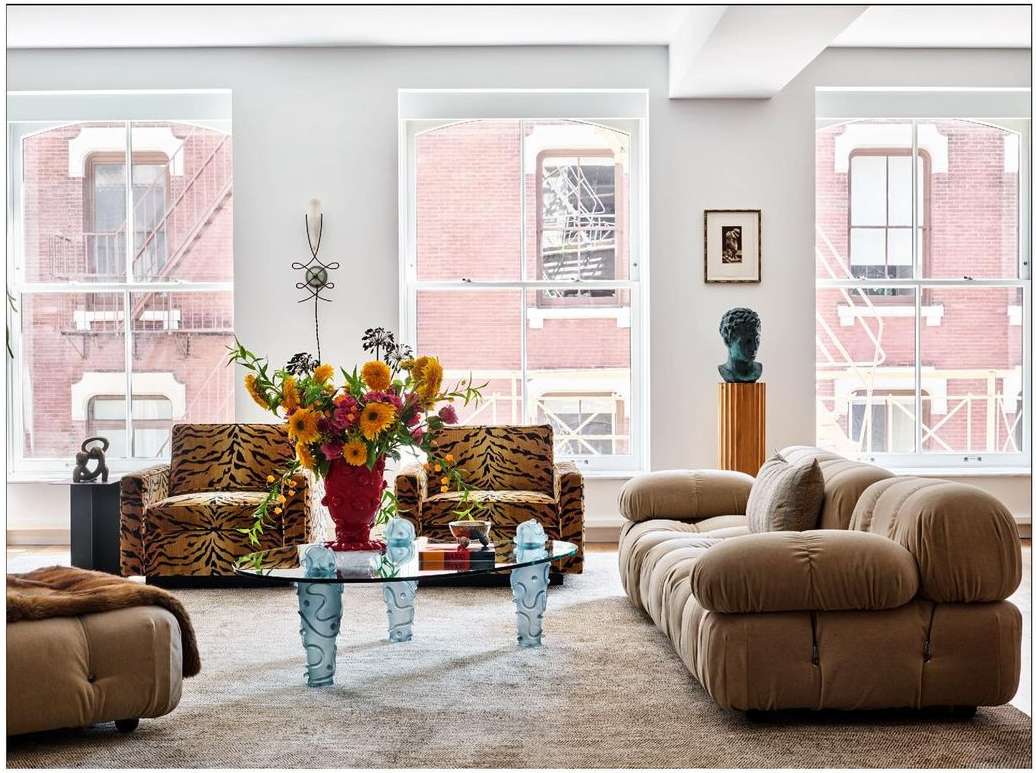 Living room interior featuring a tan sofa, patterned armchairs, glass coffee table, and flowers in a red vase against a neutral backdrop.