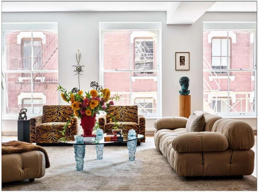 Living room interior featuring a tan sofa, patterned armchairs, glass coffee table, and flowers in a red vase against a neutral backdrop.