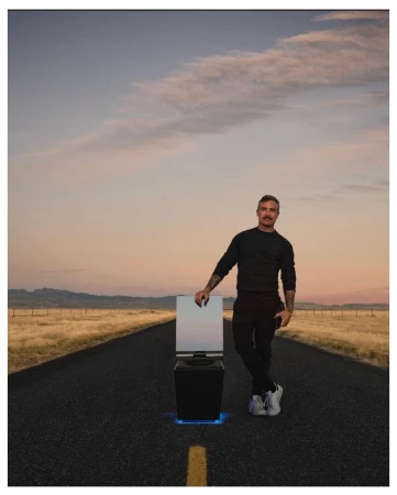 Adult male poses on a road with a smart toilet featuring a mirror. The landscape shows mountains and a golden sunset sky.