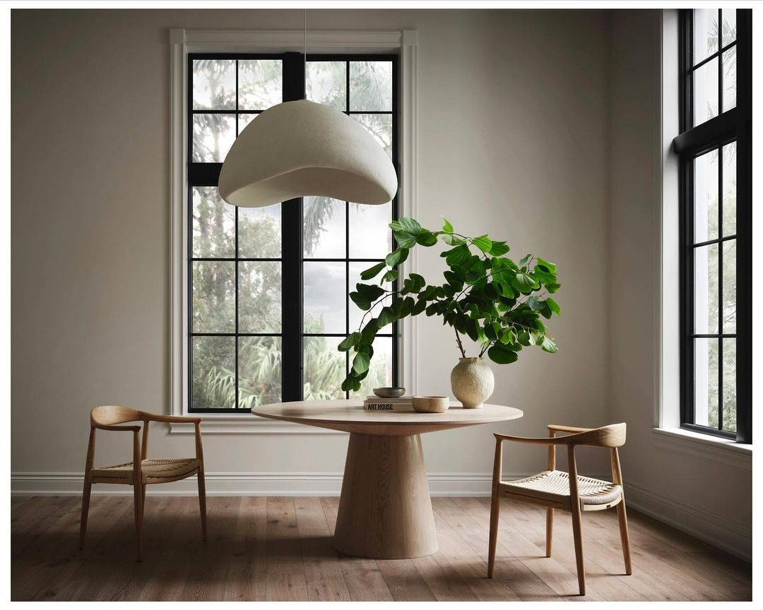 Bright indoor dining room scene with a round light wood table, vase with green leaves, modern chairs, wood floors, and natural light.