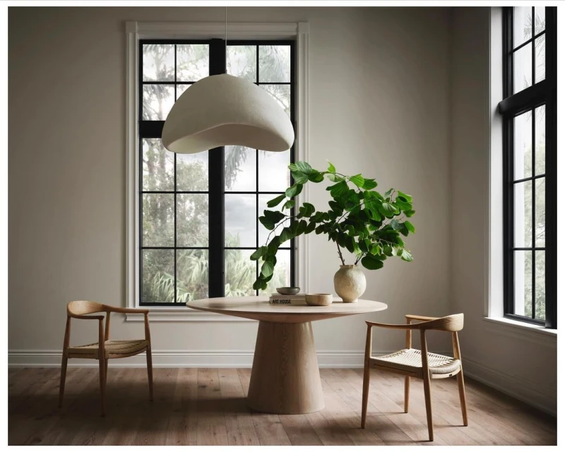 Bright indoor dining room scene with a round light wood table, vase with green leaves, modern chairs, wood floors, and natural light.