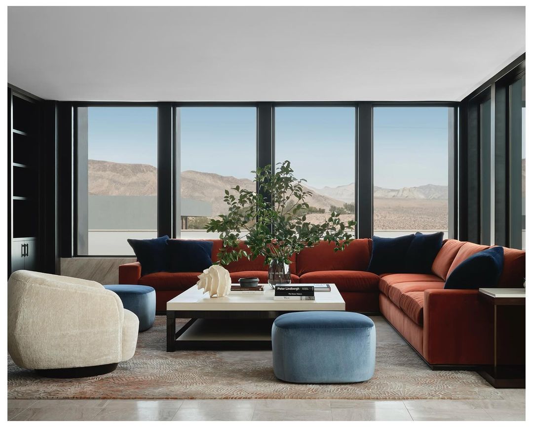 Interior of a modern living room with an orange sectional sofa, white coffee table, blue accents, and a view of desert mountains.