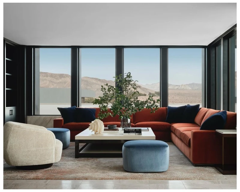 Interior of a modern living room with an orange sectional sofa, white coffee table, blue accents, and a view of desert mountains.