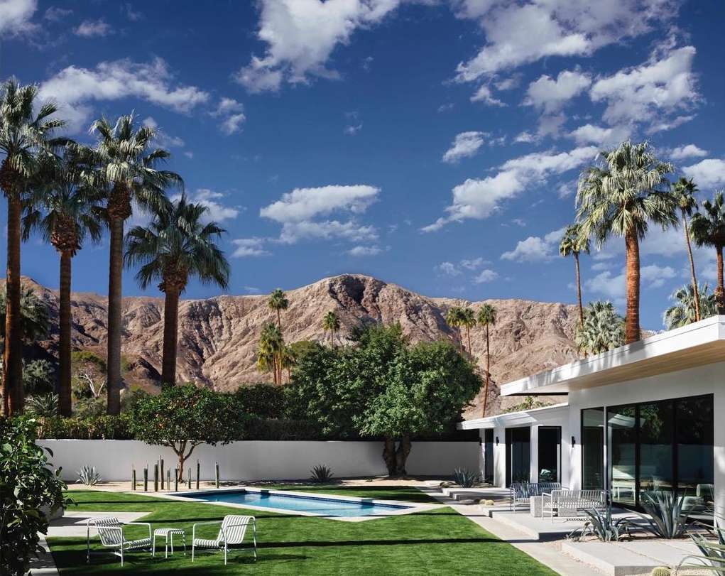 Modern house with a swimming pool and lawn in front of a mountain range, palm trees, clear blue sky with scattered clouds.