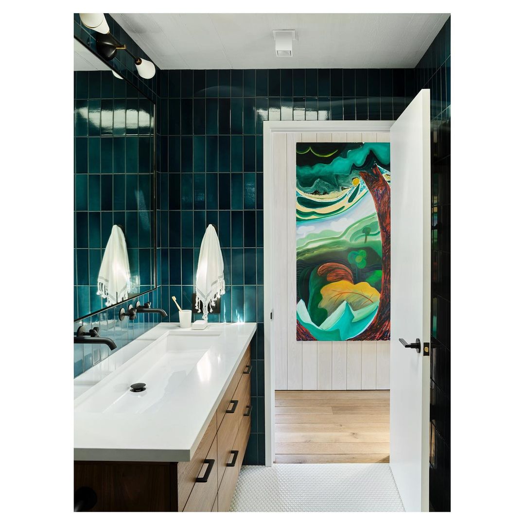 Modern bathroom interior features turquoise tiled wall, white countertop sink, artistic painting, and open doorway with bright wood floor.