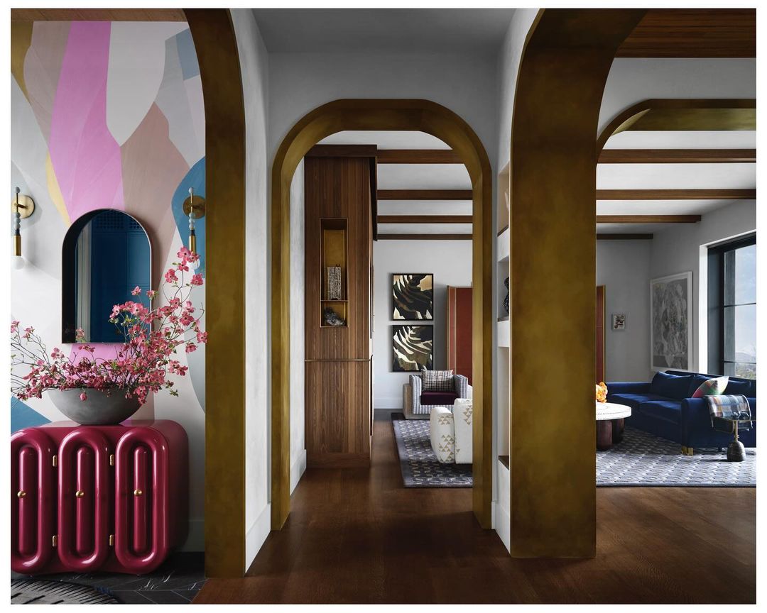 Interior view through arched doorways into a living room with a blue velvet couch, a pink credenza, and abstract wall art.