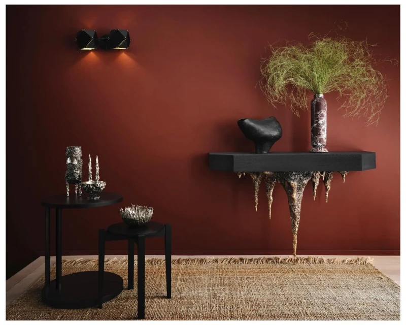 Interior view features sculptural black console table, brown vase with green foliage, with brown, textured rug in the background.