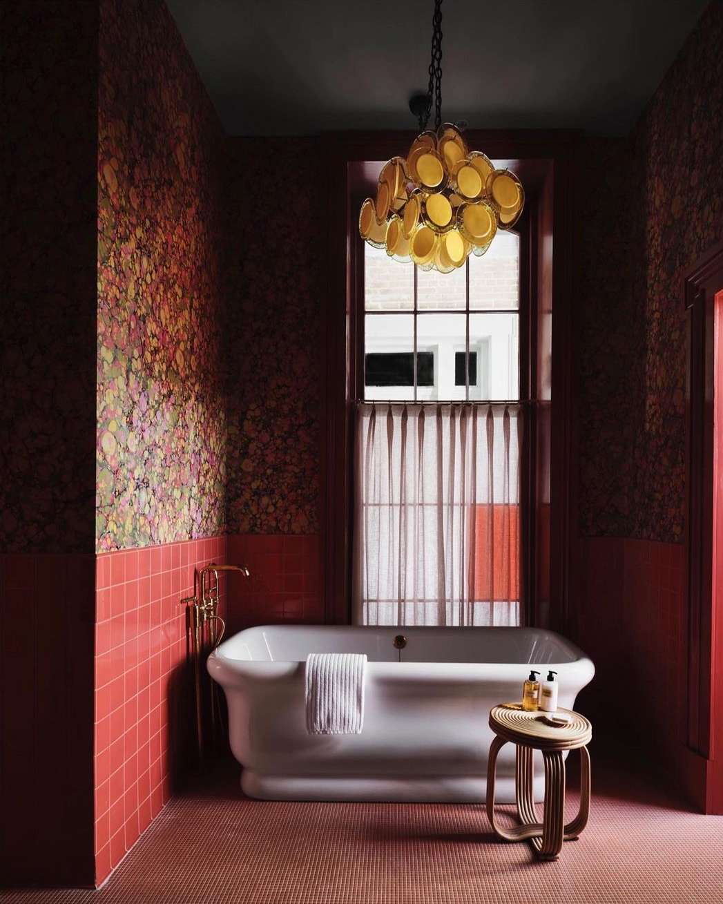 Bathroom interior featuring a white clawfoot bathtub, stool with toiletries, and a unique chandelier in a room with patterned wallpaper.
