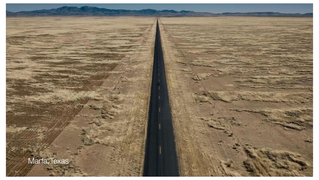 Panoramic view of a long asphalt road cutting through a vast, arid landscape with distant mountains under a clear sky, near Marfa, Texas.