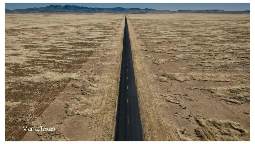 Panoramic view of a long asphalt road cutting through a vast, arid landscape with distant mountains under a clear sky, near Marfa, Texas.