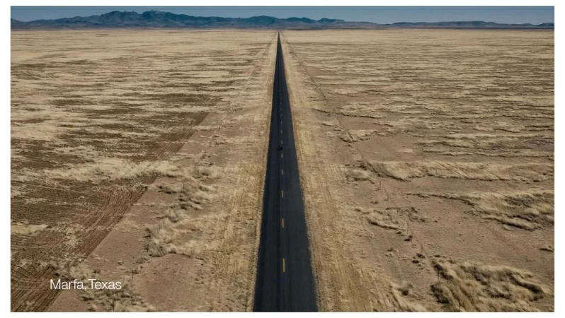 Panoramic view of a long asphalt road cutting through a vast, arid landscape with distant mountains under a clear sky, near Marfa, Texas.