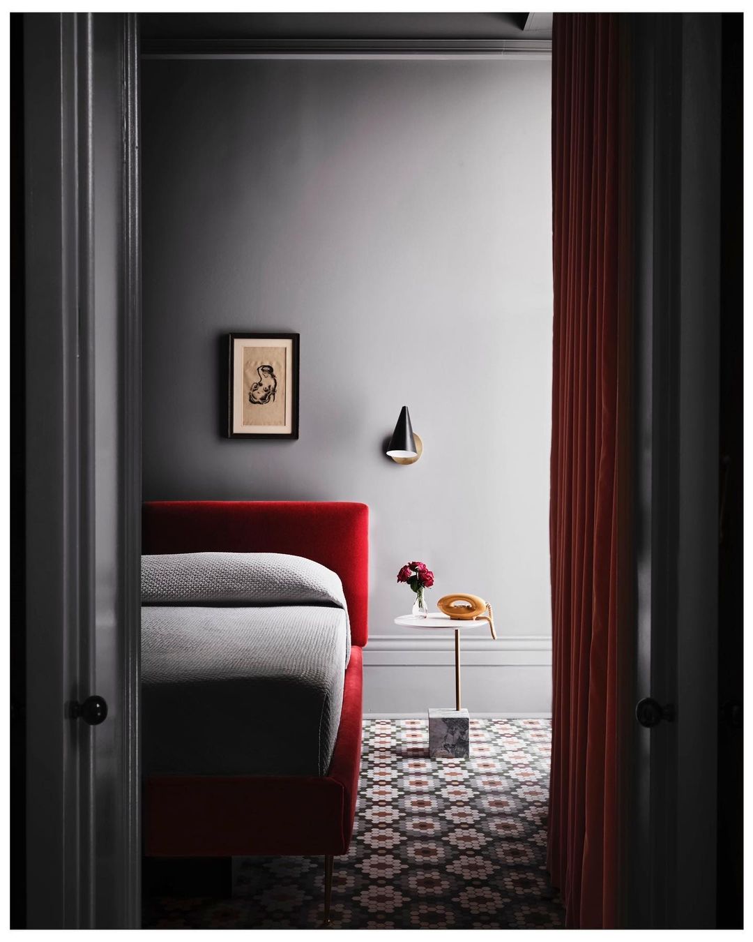 Red upholstered bed with a gray patterned floor in a minimalist bedroom with a marble-based table and a red curtain.