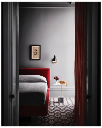 Red upholstered bed with a gray patterned floor in a minimalist bedroom with a marble-based table and a red curtain.