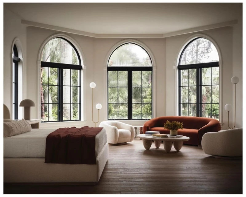 Living room interior with a bed, orange couch, cream-colored chairs, oval coffee table, globe floor lamp, and arched windows.