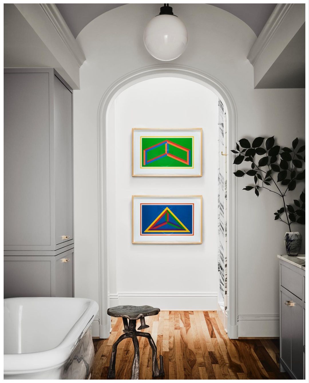 Modern bathroom interior with colorful geometric art, a freestanding tub, decorative stool, and gray cabinets in a bright, stylish space.