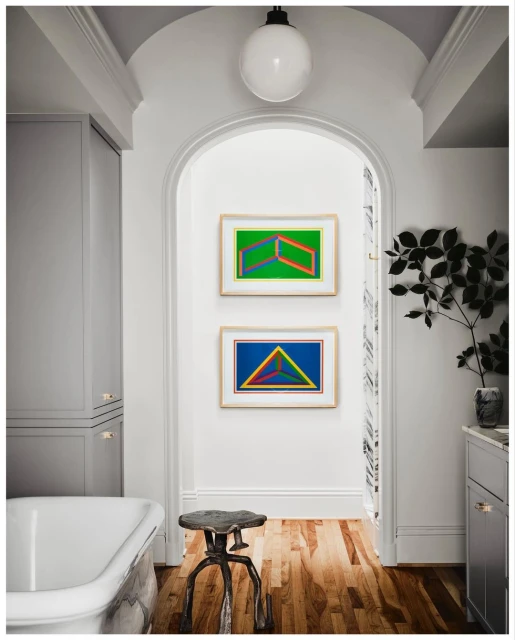 Modern bathroom interior with colorful geometric art, a freestanding tub, decorative stool, and gray cabinets in a bright, stylish space.