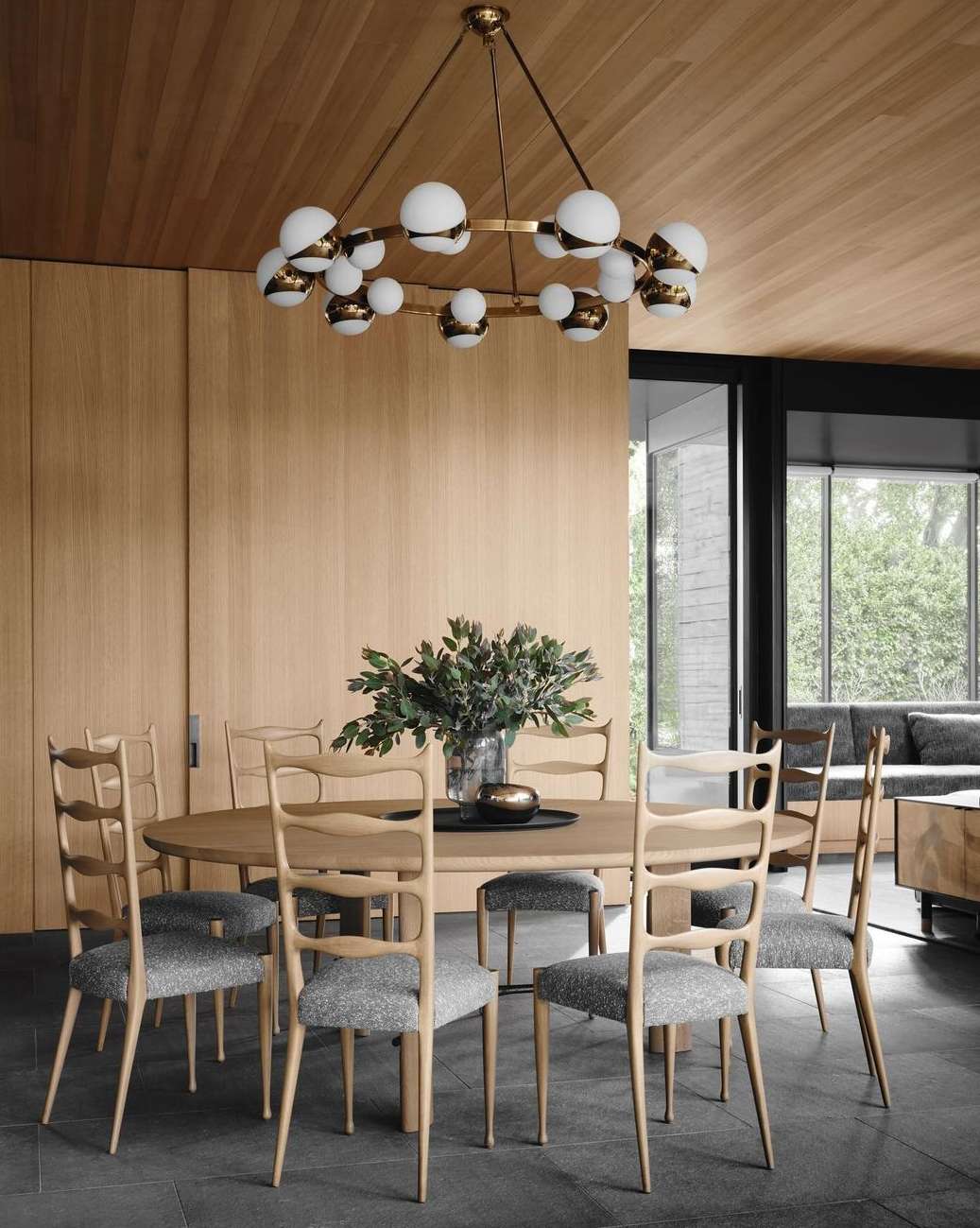 Interior of a dining room featuring a round wooden table surrounded by eight chairs under a circular chandelier with white glass globes.