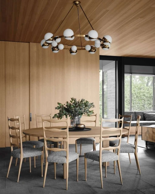 Interior of a dining room featuring a round wooden table surrounded by eight chairs under a circular chandelier with white glass globes.