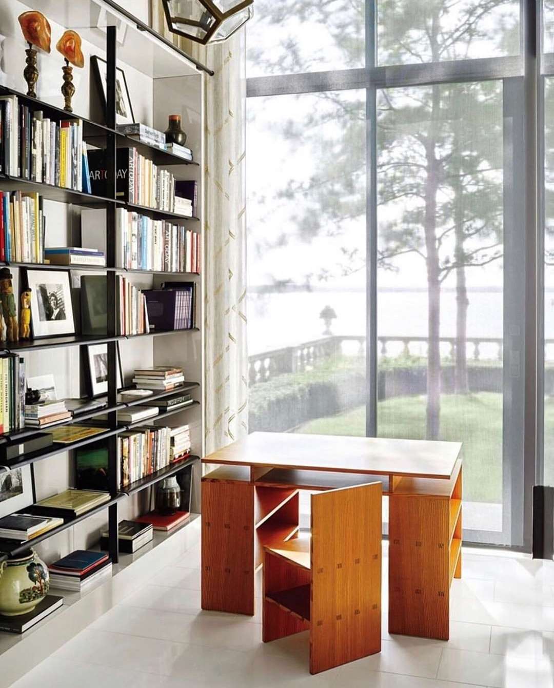 Home interior featuring a wooden table and chair set, a bookshelf filled with books and decor, and a large window with sheer curtains.