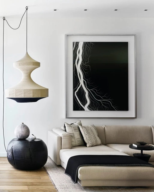 Modern interior features a beige sectional sofa, round black side table with decorative items, a hanging lamp, and framed lightning art.