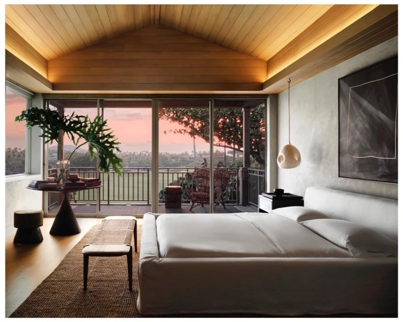 Modern bedroom interior with a large bed, woven bench, tropical plant, and sunset view from the balcony.