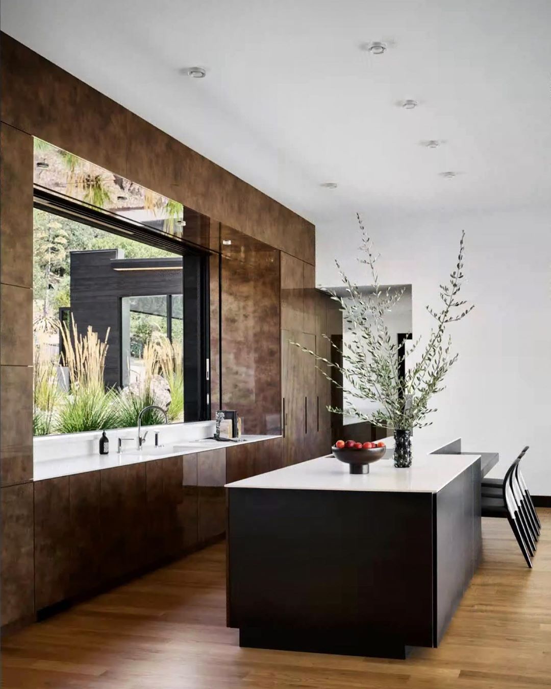 Modern kitchen interior featuring dark wood cabinets, a white countertop island, hardwood floors, and a view to an outdoor landscape.