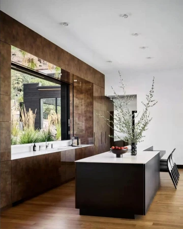 Modern kitchen interior featuring dark wood cabinets, a white countertop island, hardwood floors, and a view to an outdoor landscape.