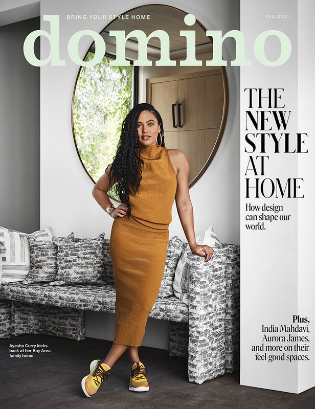 Magazine cover featuring Ayesha Curry in a mustard-colored dress and sneakers, leaning against a patterned bench with a round mirror behind.