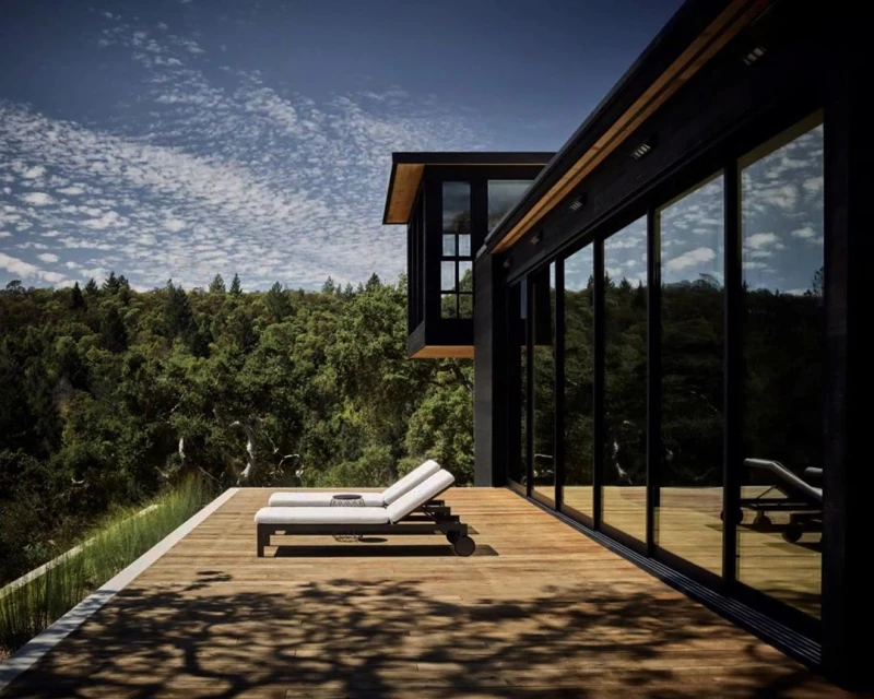 Exterior of modern home with wooden deck featuring two lounge chairs, black frame windows, and lush green trees in the background.