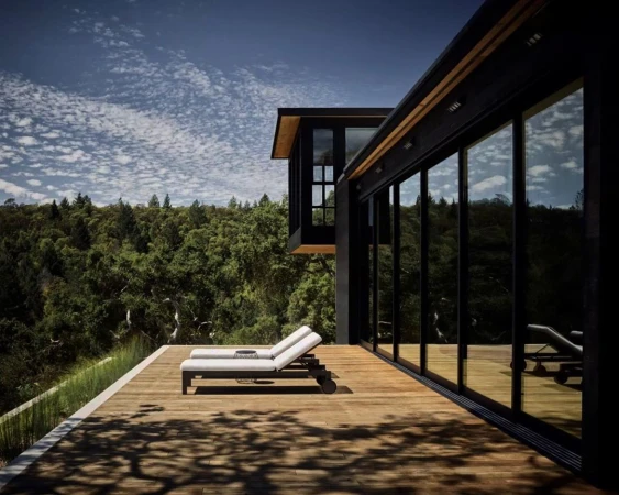 Exterior of modern home with wooden deck featuring two lounge chairs, black frame windows, and lush green trees in the background.