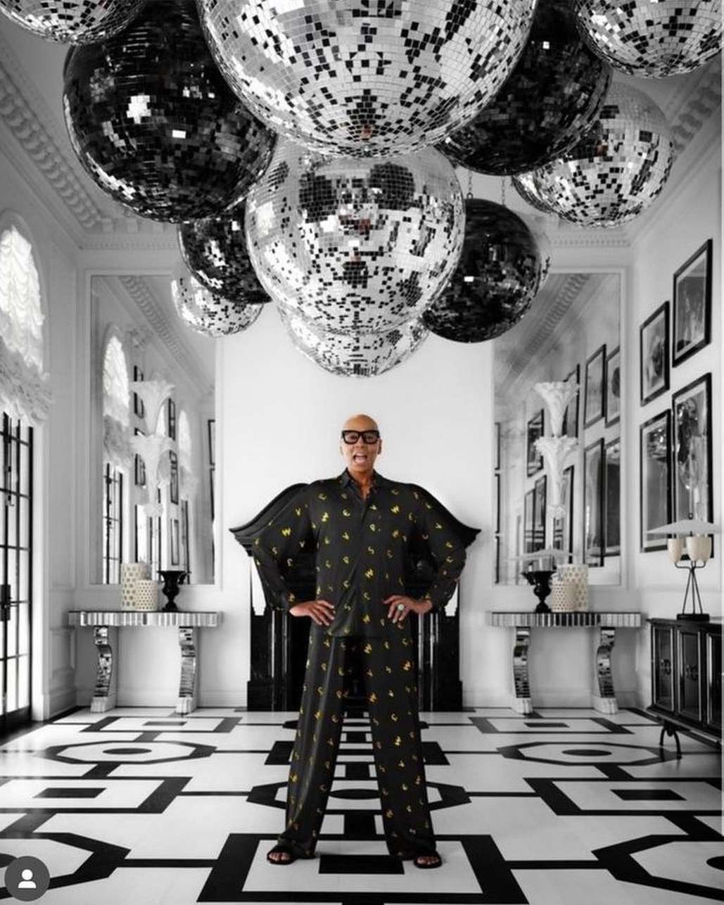 Adult male in patterned pajamas stands in a mirrored hallway with disco balls, artwork, and black-and-white patterned floor, indoor setting.