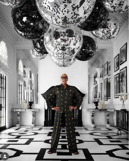Adult male in patterned pajamas stands in a mirrored hallway with disco balls, artwork, and black-and-white patterned floor, indoor setting.