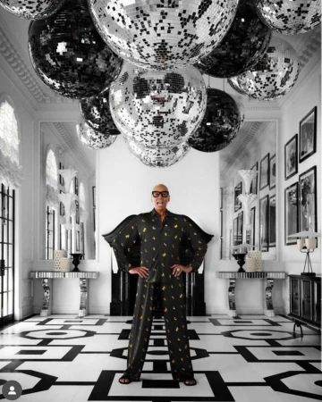 Adult male in patterned pajamas stands in a mirrored hallway with disco balls, artwork, and black-and-white patterned floor, indoor setting.