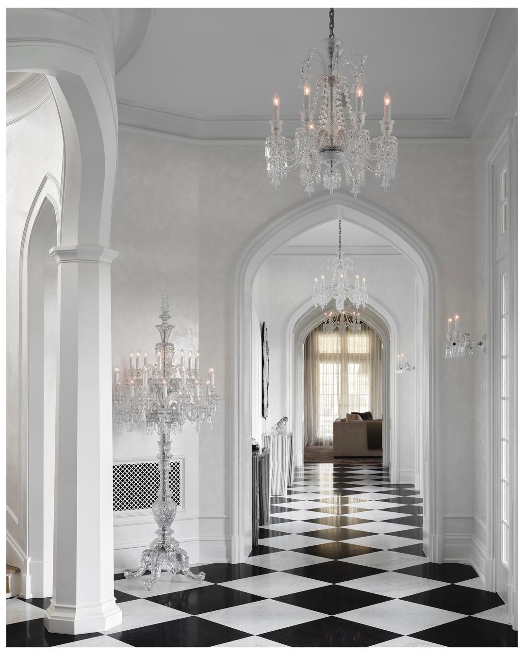 A bright, traditionally styled home foyer featuring black and white checkered tile flooring, white walls, archways, and ornate crystal chandeliers.