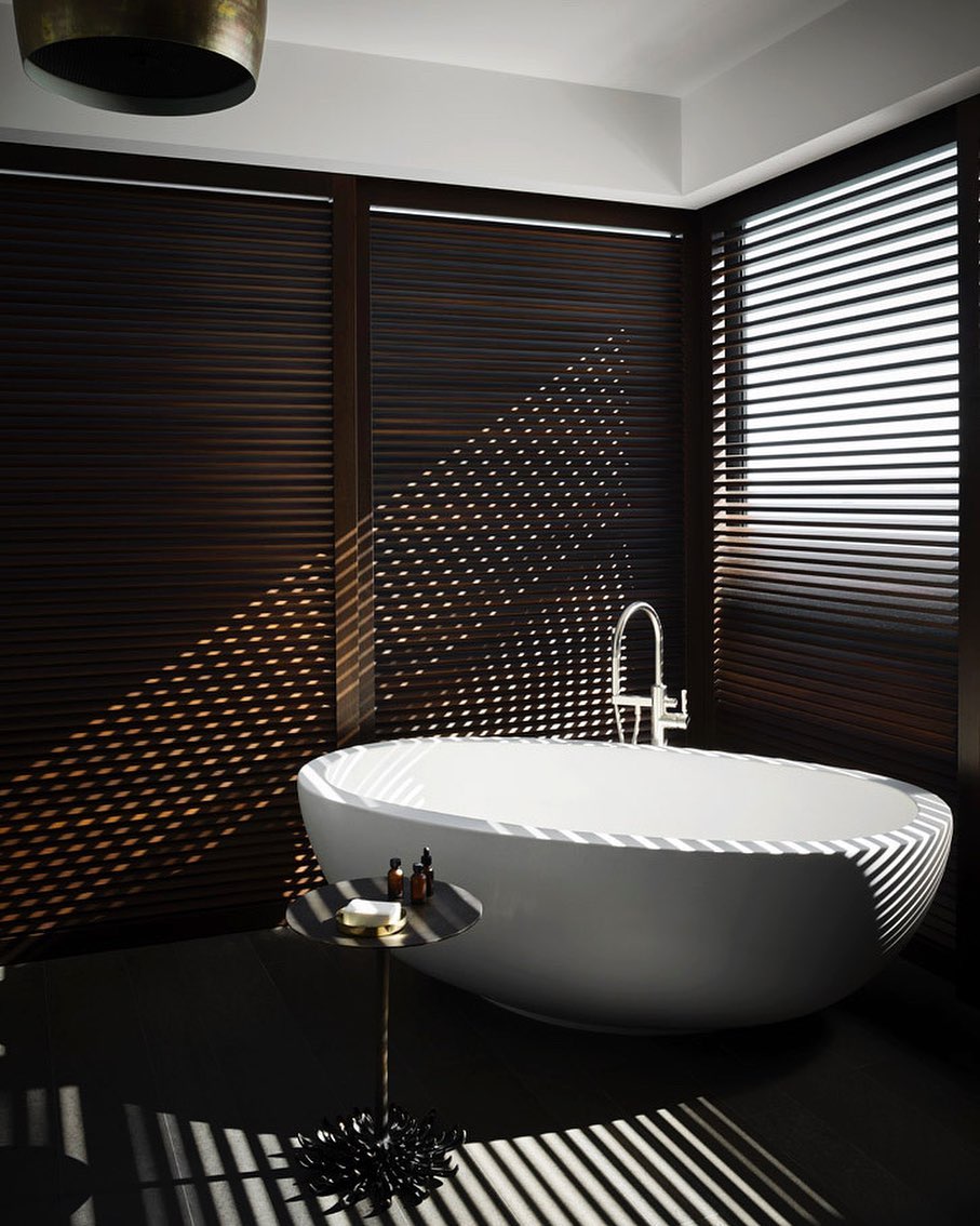 Oval freestanding bathtub in a modern bathroom with dark wood blinds, a small table with toiletries, and geometric shadows on the floor.