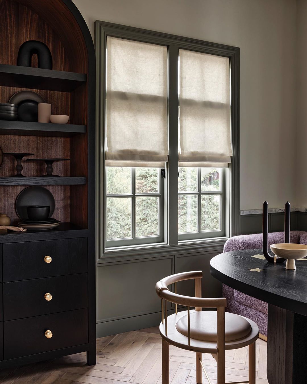 Interior shot of a home with a black cabinet, a window with beige shades, a wood chair and wood table, and hardwood floors.
