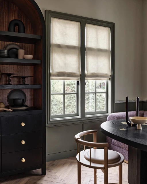 Interior shot of a home with a black cabinet, a window with beige shades, a wood chair and wood table, and hardwood floors.