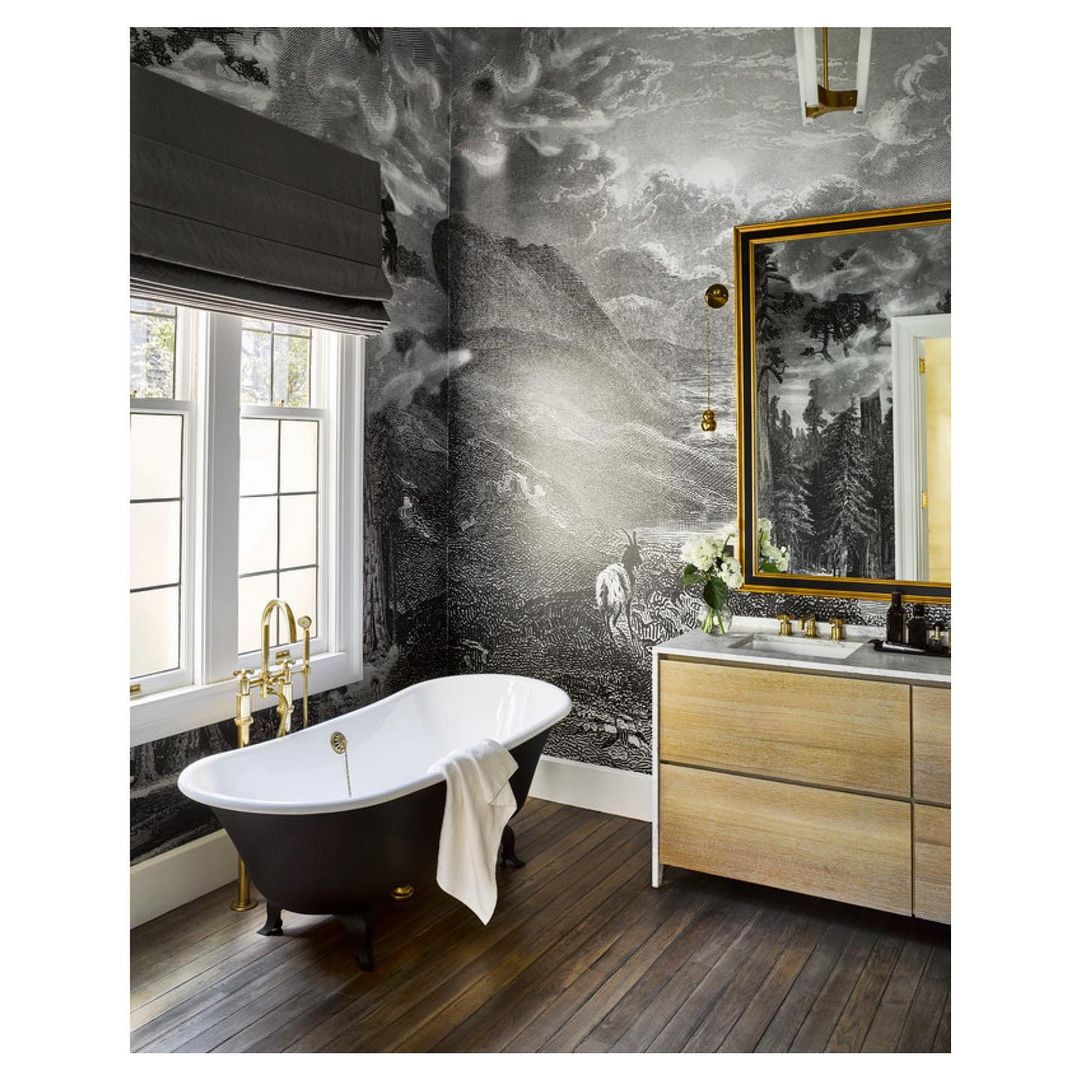 Interior of bathroom with freestanding tub, wood vanity, landscape wallpaper, framed mirror, and natural light from a window.
